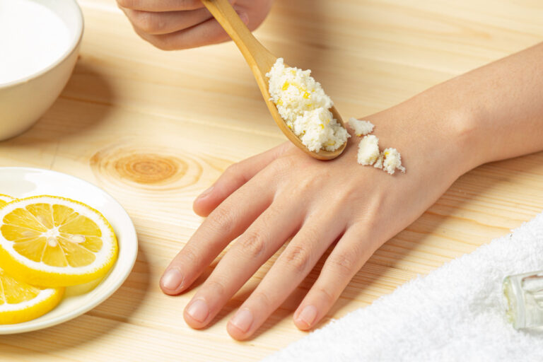 A hand on a wooden surface having a homemade scrub applied to it with a spoon, with lemon slices nearby.